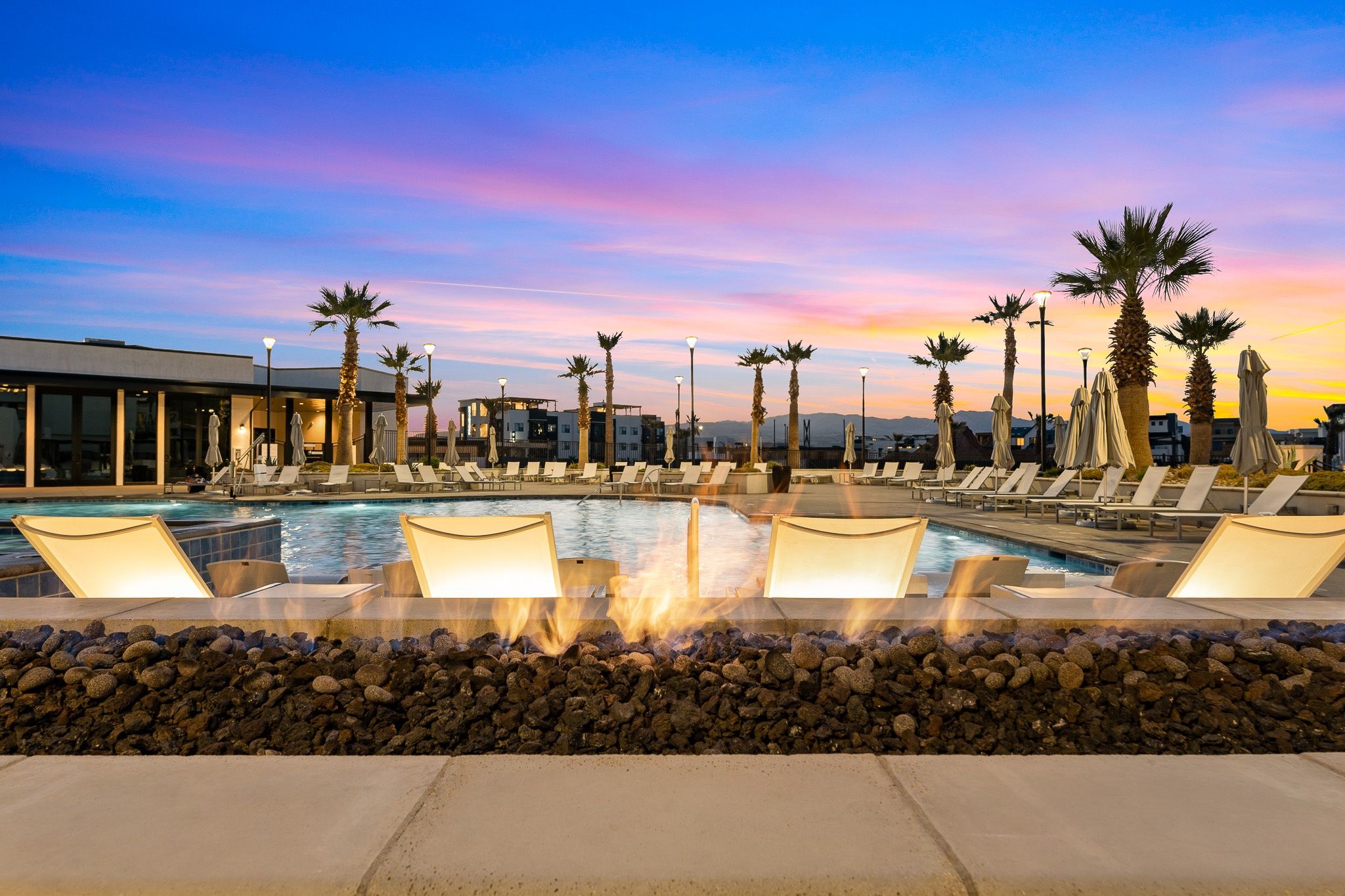 A pool area with lounge chairs and a fire feature, surrounded by palm trees and a modern building.