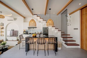 A modern kitchen with a central island, bar stools, and a staircase with wooden steps and black railings. - Villa Scorpia 4 Bingin (Pecatu)