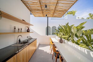 A modern kitchen with a built-in sink, wooden cabinets, and a white countertop. There is a dining area with a long table and chairs, and a pergola providing shade. - Villa Scorpia 4 Bingin (Pecatu)
