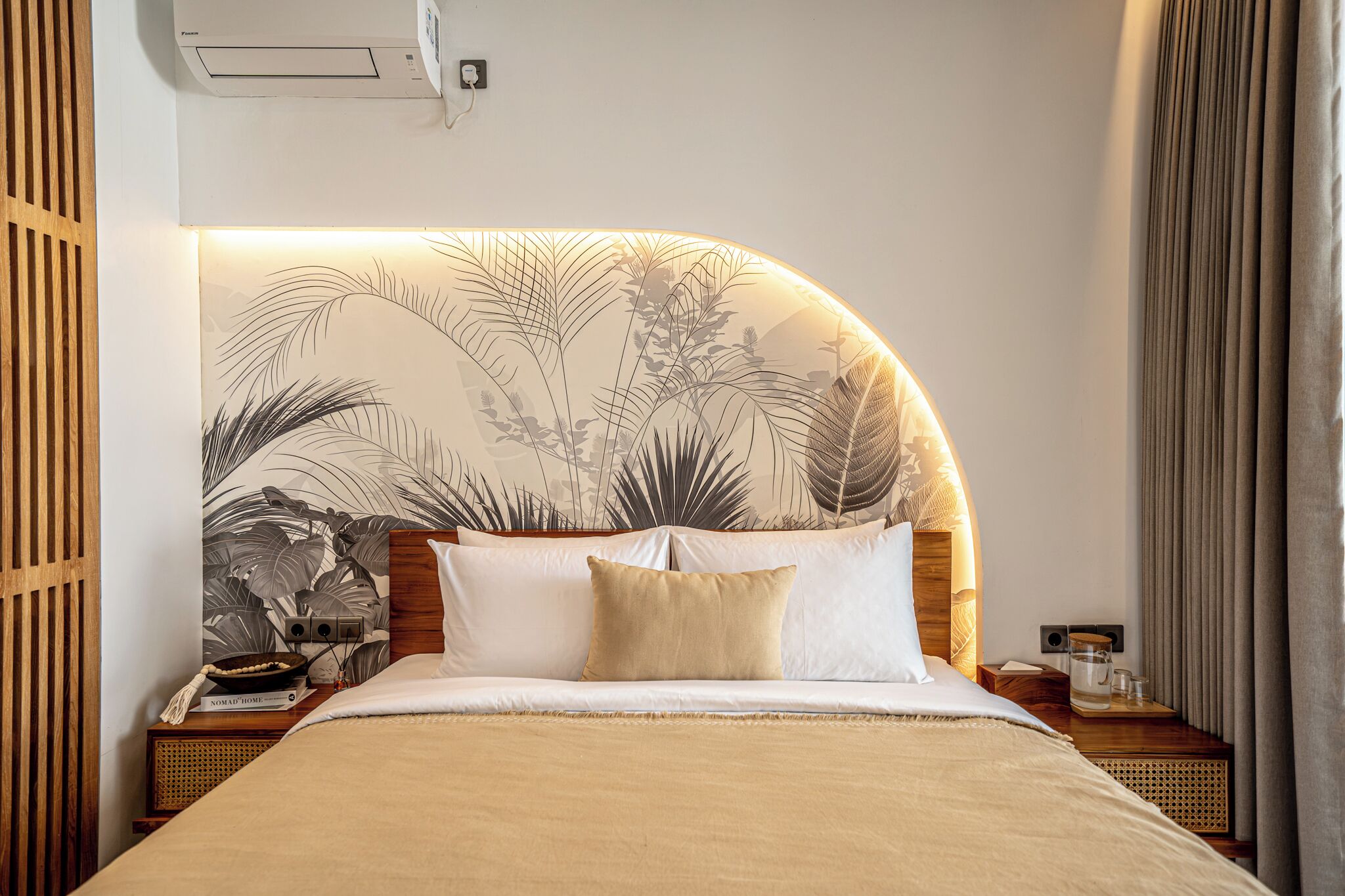 Une chambre d’hôtel avec un lit, une tête de lit ornée d’un motif de feuilles, une table de chevet avec une lampe et une fenêtre avec des rideaux.