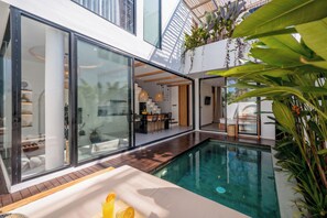A modern outdoor pool area with glass walls, a dining area, and lush greenery. - Villa Scorpia 4 Bingin (Pecatu)