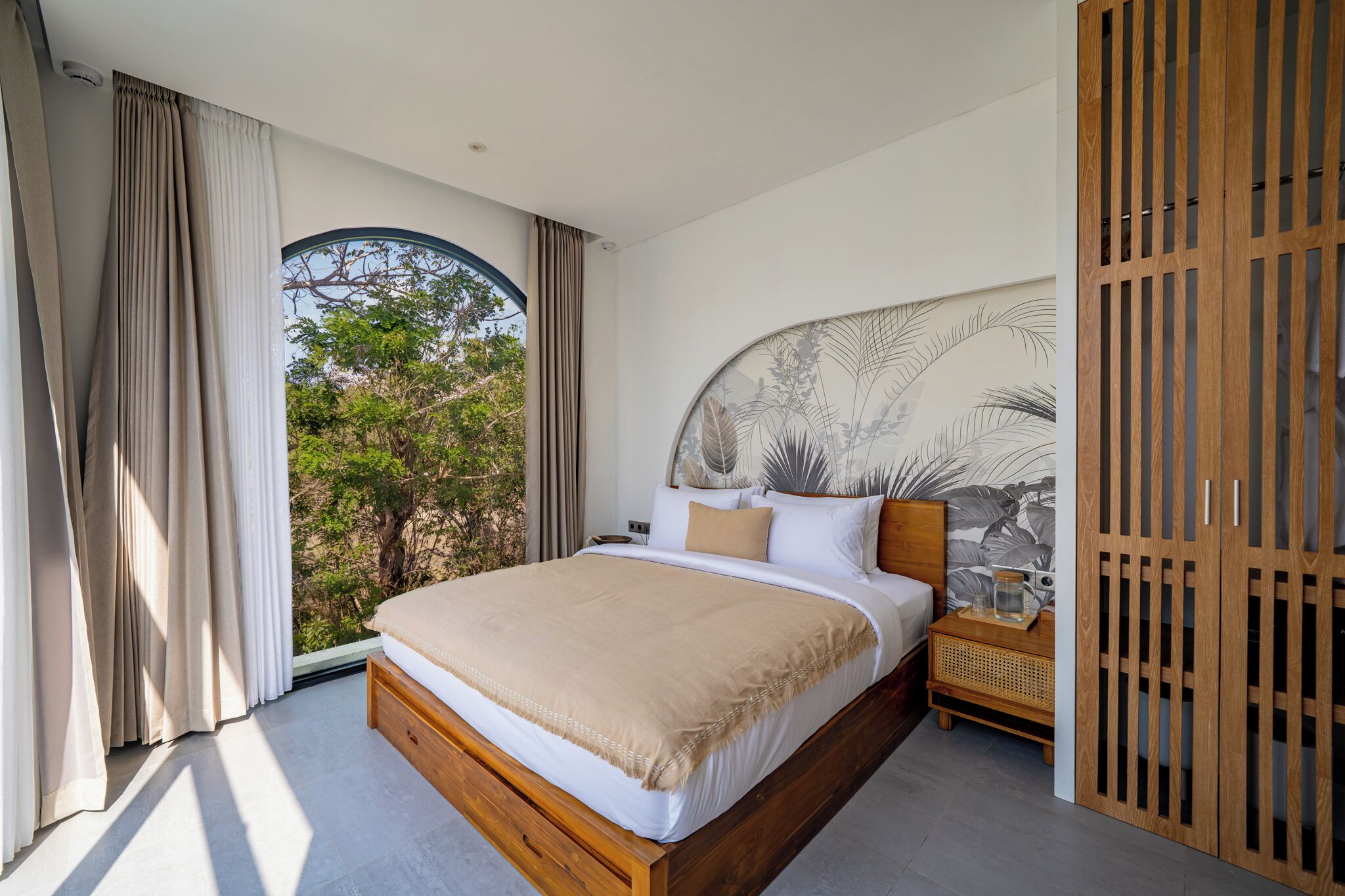 A bedroom with a large bed, a nightstand, a wall-mounted shelf, and a view of trees through a window.