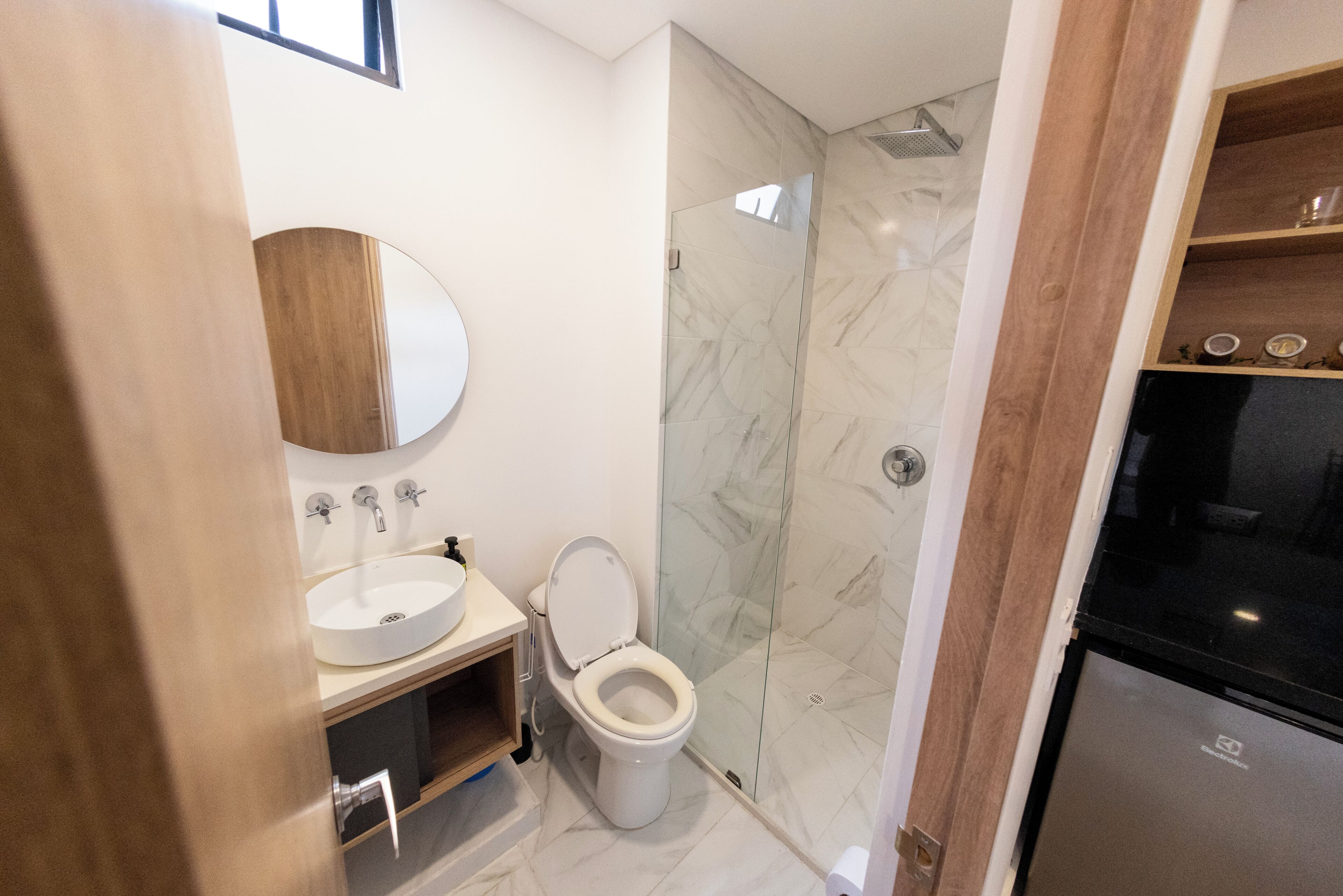 Un baño moderno con lavamanos, espejo y retrete blancos, una cabina de ducha de vidrio y un refrigerador negro.