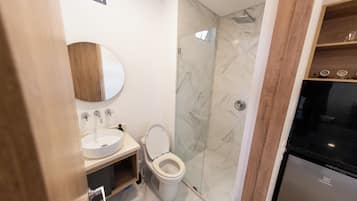 Un baño moderno con lavamanos, espejo y retrete blancos, una cabina de ducha de vidrio y un refrigerador negro.