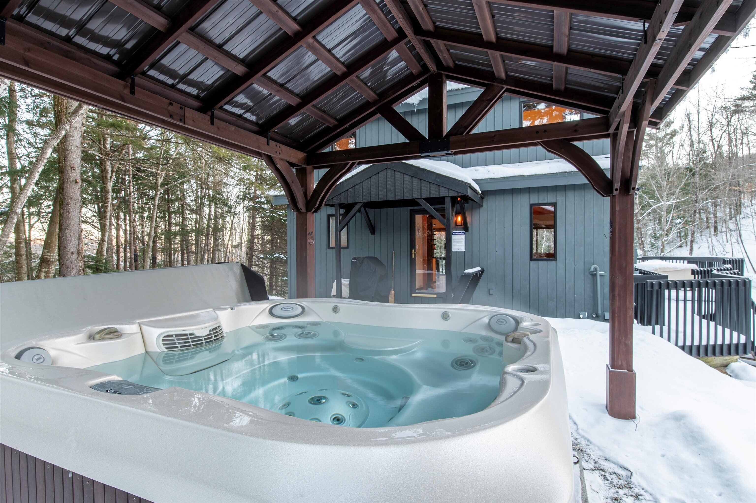 A hot tub under a wooden pergola with a view of a cabin and trees.