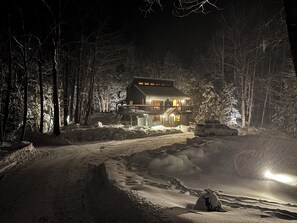 A snow-covered landscape with a house featuring a balcony and a driveway leading to a vehicle. - Okemo Ski House - Hot Tub, Fireplace, Sleeps 10 - Secluded house with privacy (Ludlow)