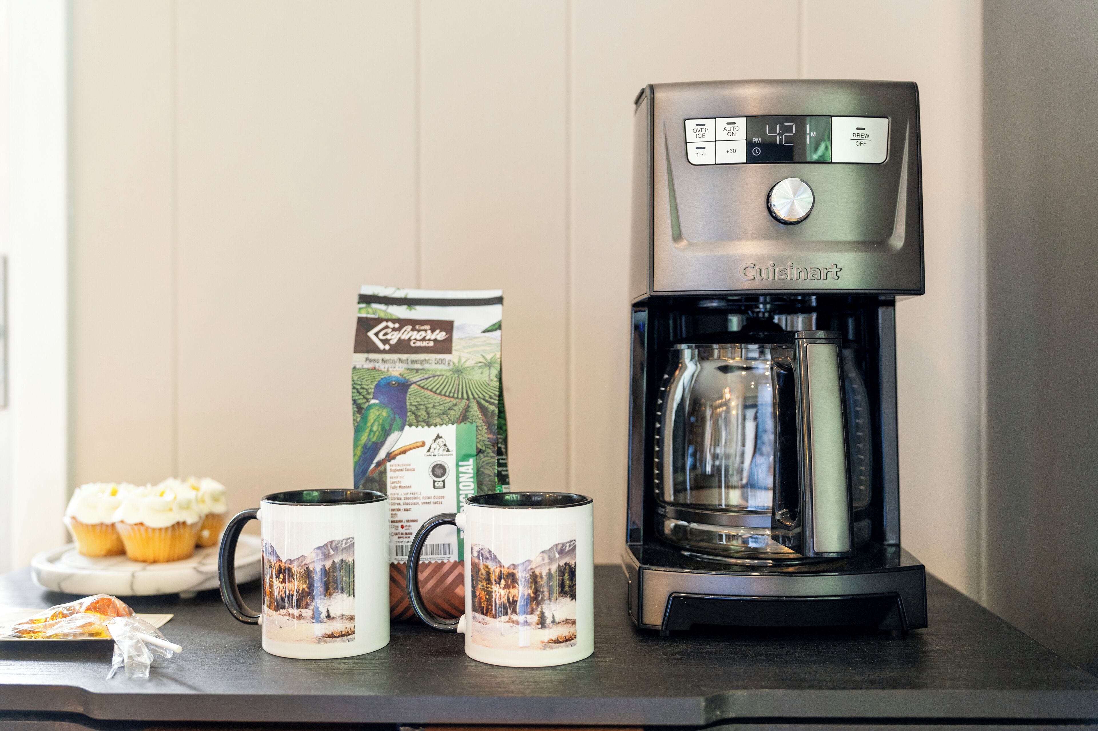 Una cafetera con pantalla digital, dos tazas con diseño escénico y una caja de cápsulas de café sobre una encimera de cocina.