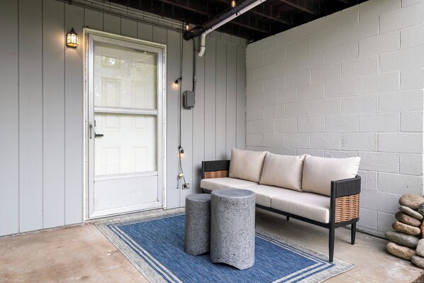 A covered porch with a white door, a light fixture, a sofa with cushions, a round stone table, and a blue rug.