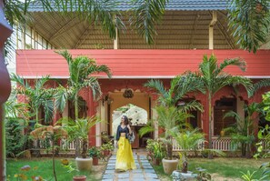 A covered outdoor area with a red building, potted plants, and a pathway. - Eartham Stays Pushkar (Pushkar)