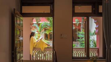 Una persona leyendo un libro en una habitación con balcón, una puerta de madera y vistas a un edificio rojo y vegetación.