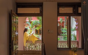 Uma pessoa lendo um livro em um quarto com varanda, porta de madeira e vista para um prédio vermelho e vegetação.