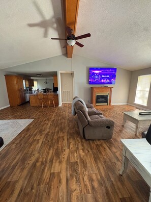 A spacious living area with a wooden floor, a reclining sofa, a television mounted above a fireplace, and a kitchen in the background. -  Can hunt on TVA land  Hunters and Fishermans Guntersville Getaway   3BR • 2BA (Langston)