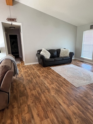 Sala de estar com sofá de couro escuro, tapete claro e piso de madeira.
