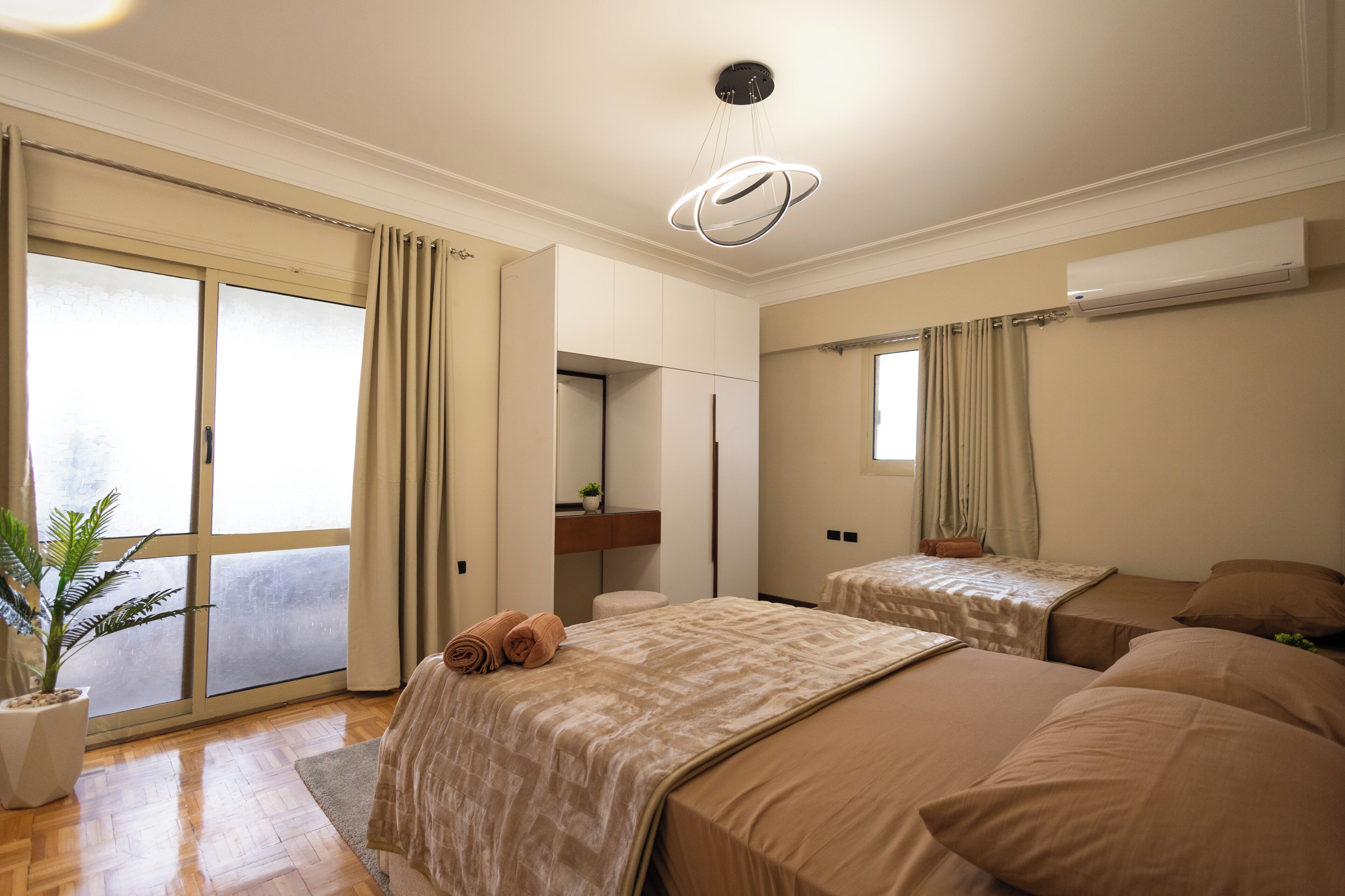 Habitación de hotel con un gran ventanal corredero, una cama con una manta color crema, una mesita de noche pequeña con una planta y una lámpara de techo.