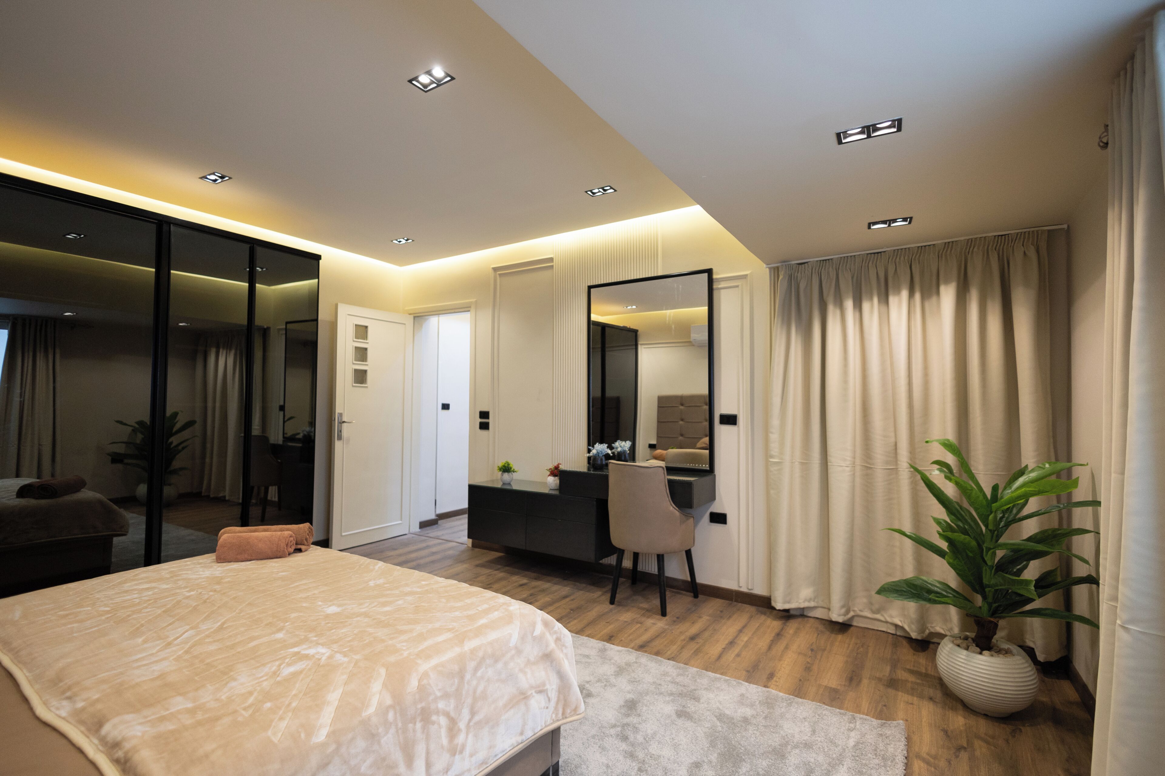 A modern bedroom with a bed, a desk, a chair, a large mirror, and a potted plant.