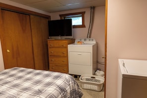 Un dormitorio con cama, cómoda, televisor, lavadora y cesto de ropa.