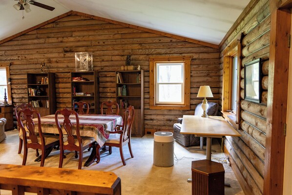 A log cabin interior with a dining table, chairs, bookshelves, a lamp, and a window.