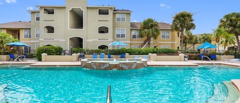 A swimming pool with a fountain, surrounded by lounge chairs and umbrellas, with a multi-story building in the background.