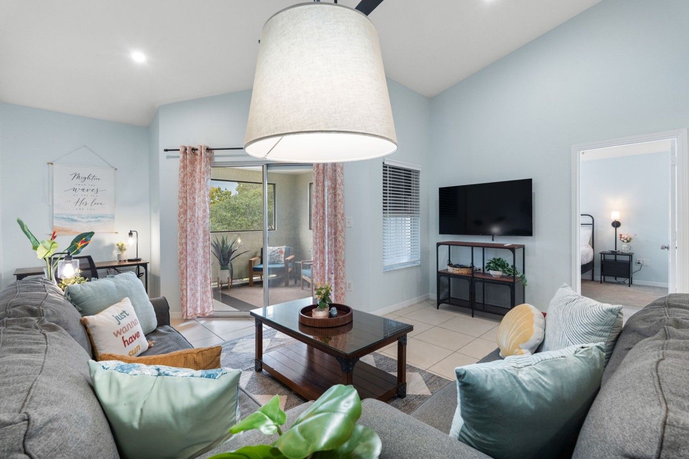 A modern living room with a sofa, a coffee table, and a television. There is a sliding glass door leading to a balcony.