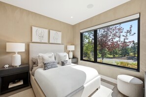 Ein modernes Schlafzimmer mit einem großen Bett, zwei Nachttischlampen und Blick auf Bäume durch ein großes Fenster.
