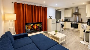 A modern living room with a sofa, a flat-screen TV, a coffee table, and a kitchen area with appliances. - Wimbledon Lodge (London)