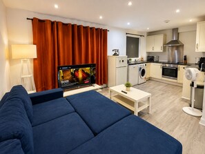 A modern living room with a sofa, a flat-screen TV, a coffee table, and a kitchen area with appliances. - Wimbledon Lodge (London)