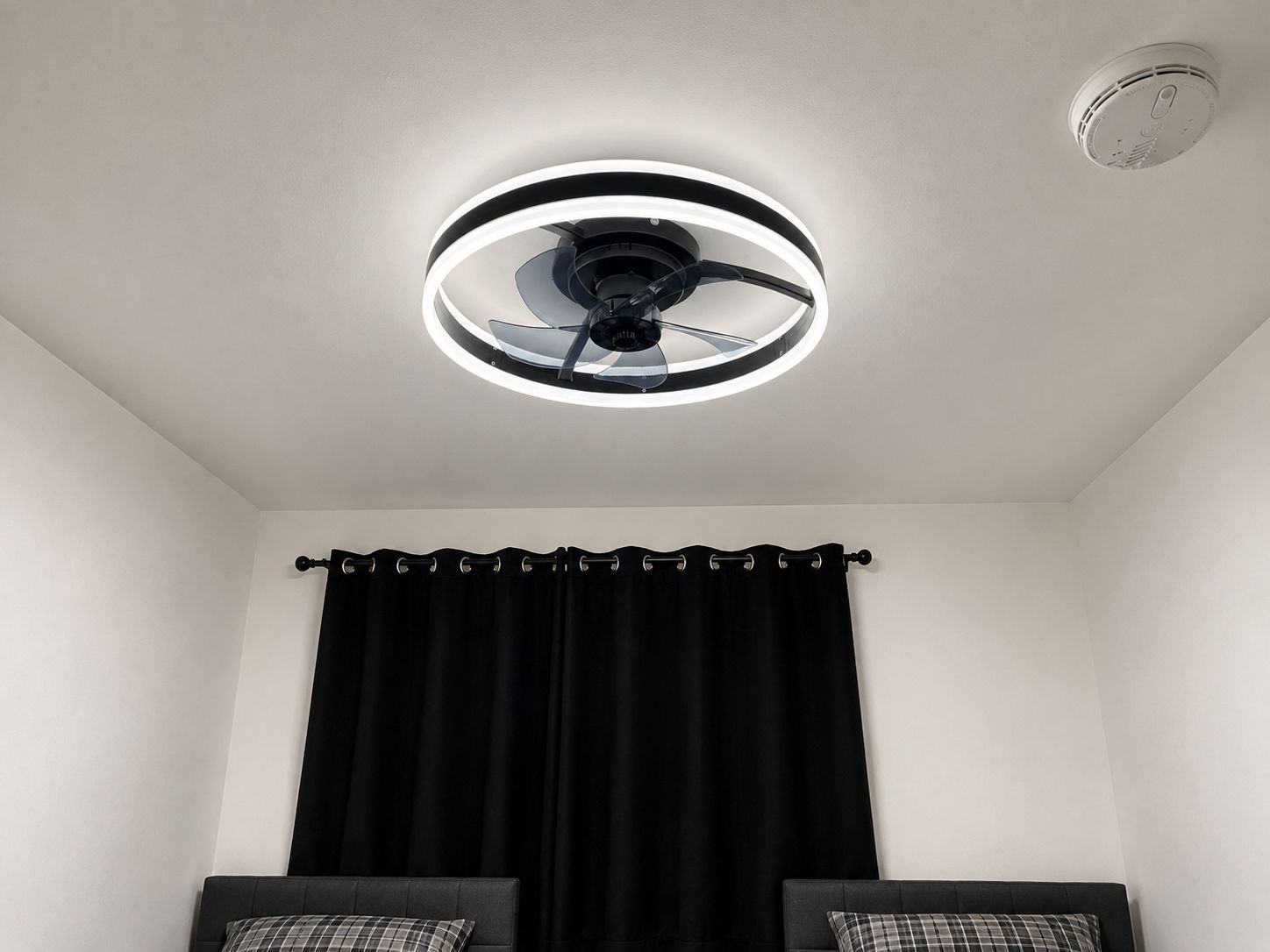 Un ventilador de techo con lámpara circular, una cortina negra y una cama con cabecera.