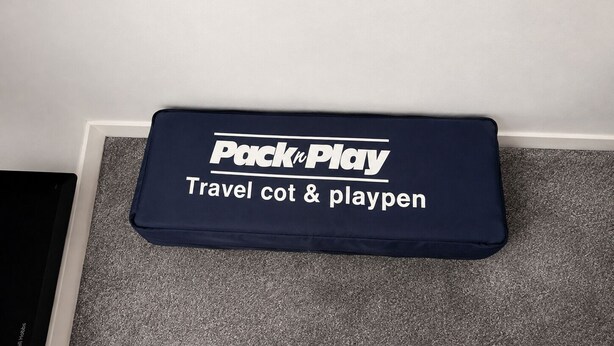 A dark blue padded bench with 'Pack'n Play' branding and text 'Travel cot & playpen' on a carpeted floor. - Wimbledon Lodge (London)