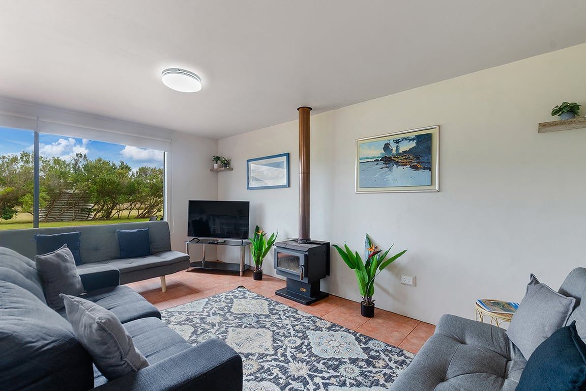 A modern living room with a sofa set, a television, a wood-burning stove, and a large window offering a view of greenery.