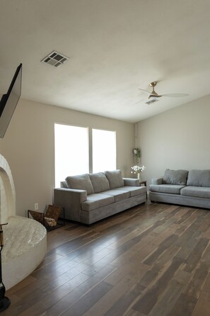 Ein geräumiges Wohnzimmer mit zwei grauen Sofas, einem Deckenventilator und großen Fenstern.