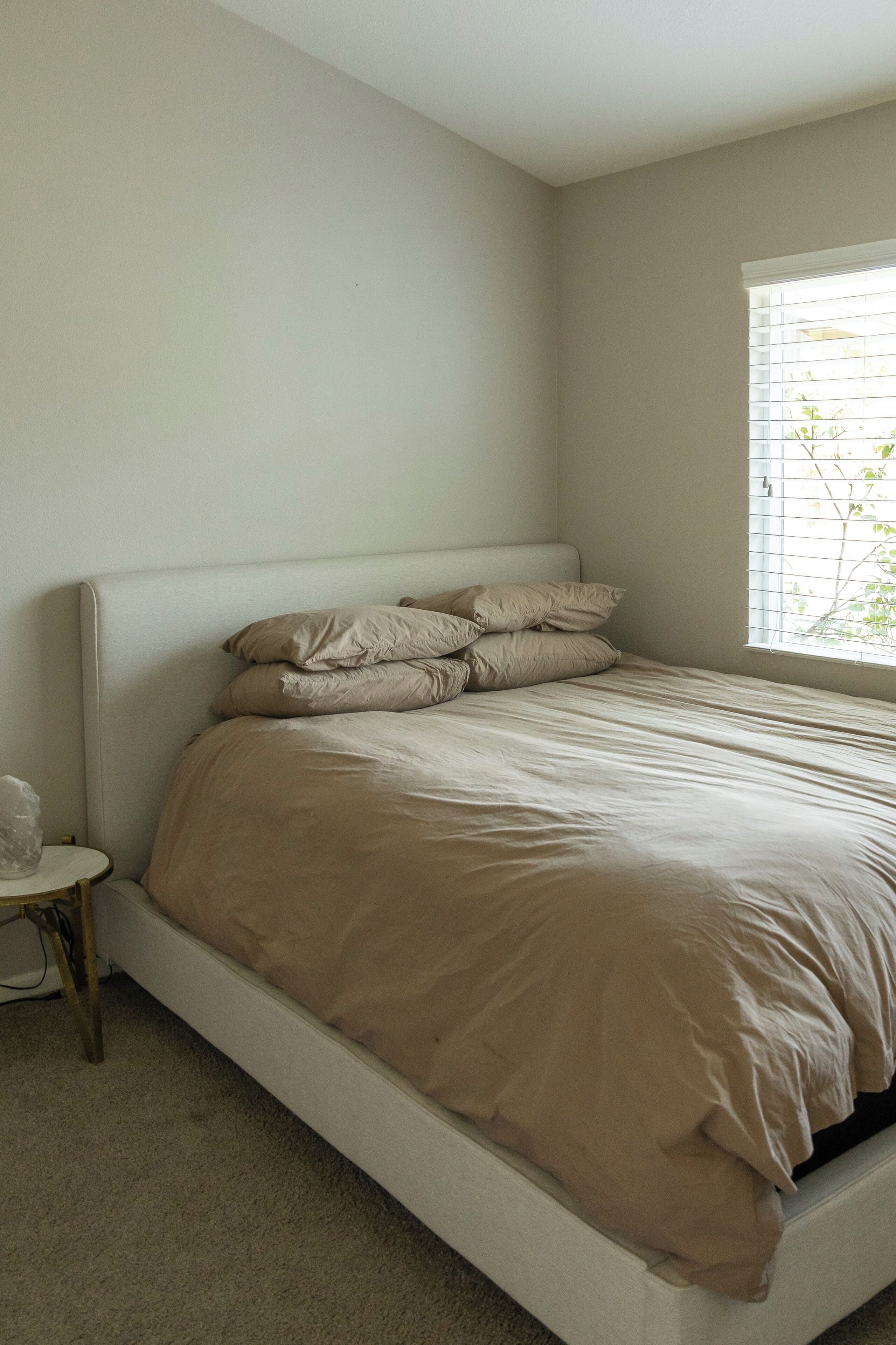 Habitación individual con una cama de una sola plaza, cabecera blanca, ropa de cama color beige, una mesita de noche pequeña y una ventana con persianas.