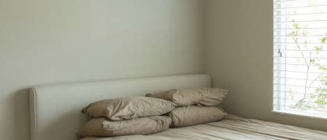 Habitación individual con una cama de una sola plaza, cabecera blanca, ropa de cama color beige, una mesita de noche pequeña y una ventana con persianas.