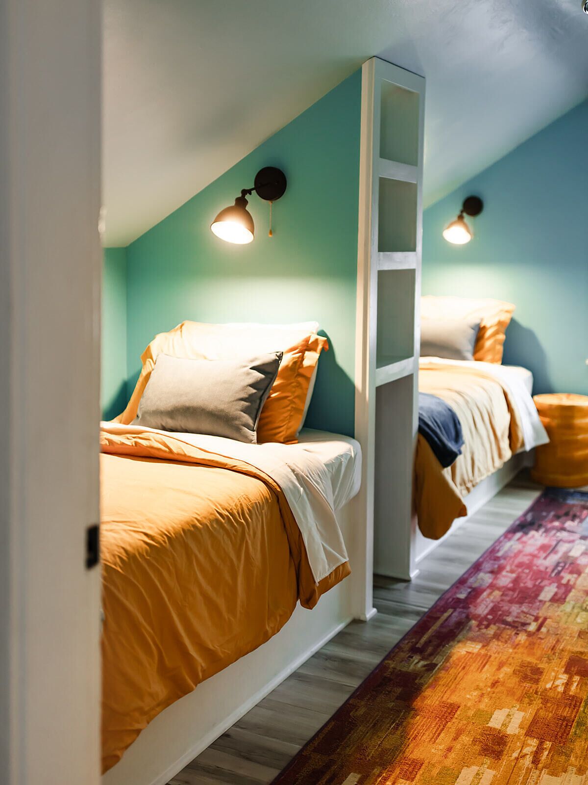 Una habitación con dos camas, cada una con cabecera blanca y ropa de cama naranja. Hay dos lámparas de pared a cada lado de las camas.