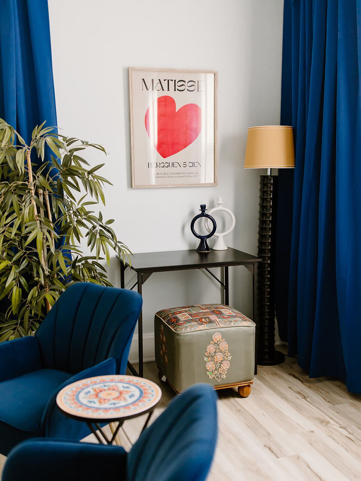 Una habitación con una silla azul, una mesita redonda pequeña, una lámpara, una obra de arte enmarcada y una planta en maceta.