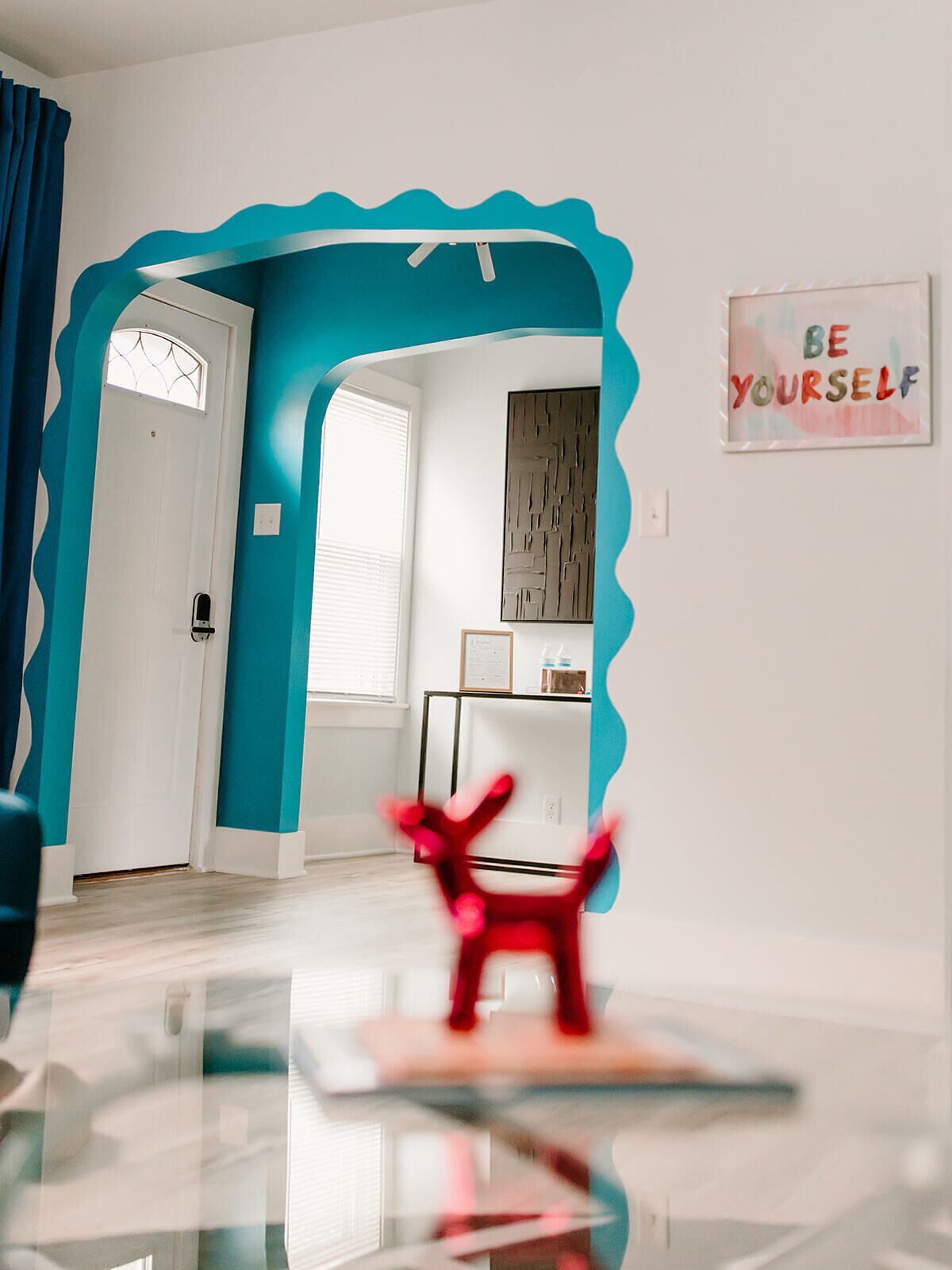 Ein Zimmer mit einer türkisfarbenen Akzentwand, einer weißen Tür, einem Fenster mit Jalousien, einem gerahmten Kunstwerk und einem roten Sessel.