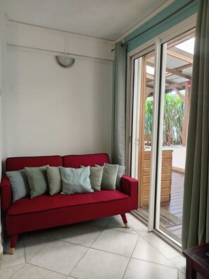 A red sofa with multiple cushions, a tiled floor, and a sliding glass door leading to an outdoor area with wooden flooring and greenery. - 2-bedroom apartment in peaceful Les Trois-Îlets with AC (Les Trois-Îlets)