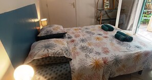 Un lit recouvert d’une couvre-lit à motifs floraux, deux serviettes enroulées, une lampe de chevet et une vue sur un espace extérieur grâce à une porte vitrée.