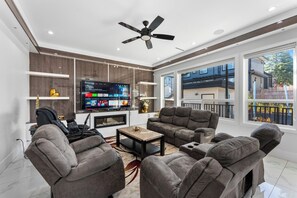 A modern living room with a flat-screen TV, grey sofas, a ceiling fan, and large windows. - Grand 5BR Luxury Home ! AC and Home Theater (Surrey)