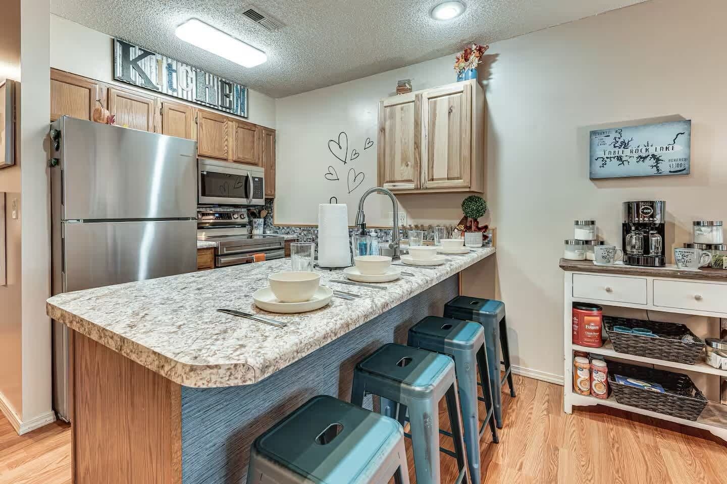 A modern kitchen with a granite countertop, stainless steel appliances, wooden cabinets, and bar stools.