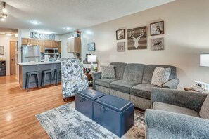 Ein modernes Wohnzimmer mit einem grauen Sofa, passendem Hocker und einem gemusterten Sessel. Im Hintergrund ist eine Küche mit Holzausbau und einer Bar erkennbar.
