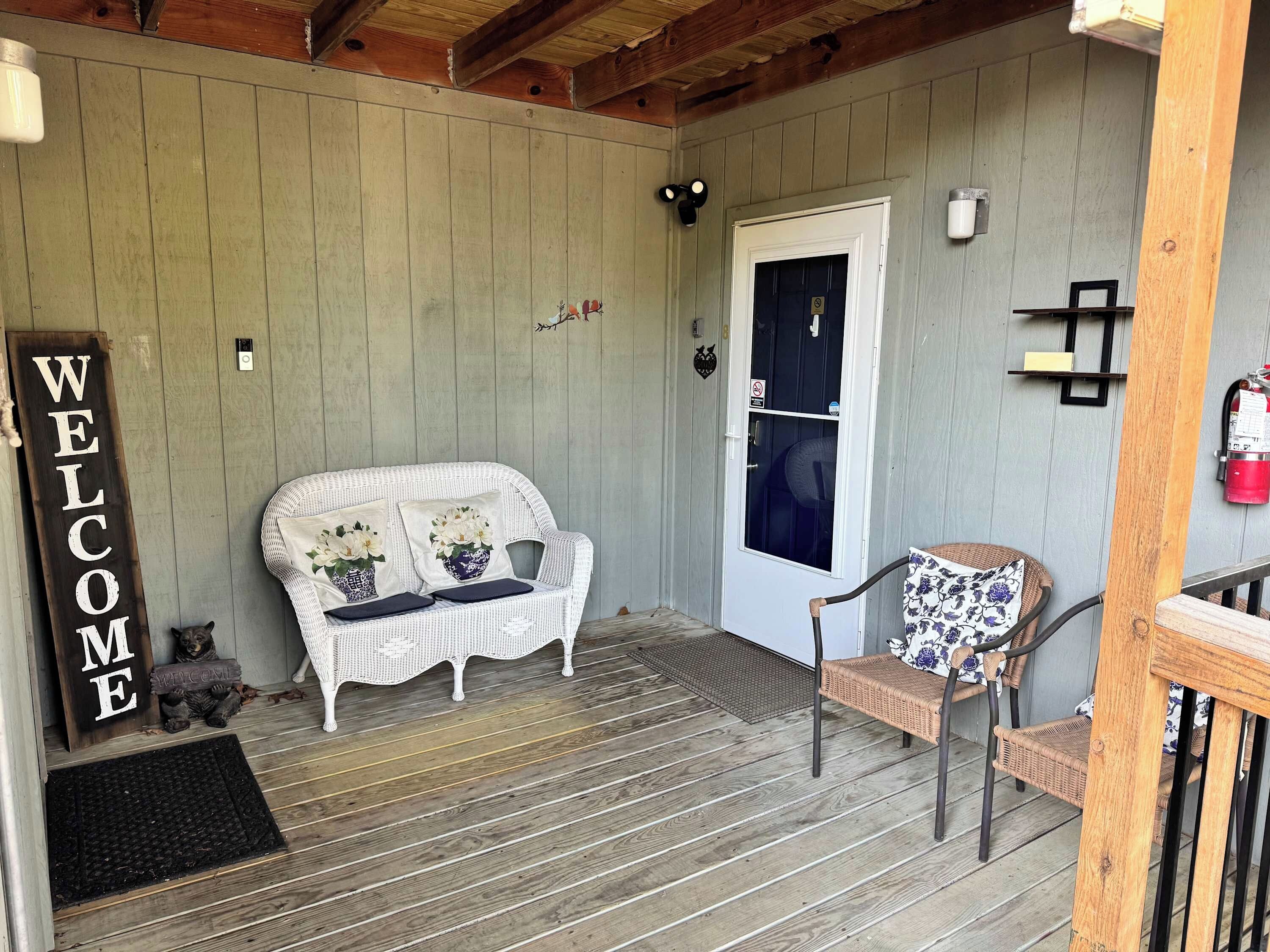A porch with a white wicker sofa, a wicker chair, a welcome sign, a door, and a fire extinguisher.