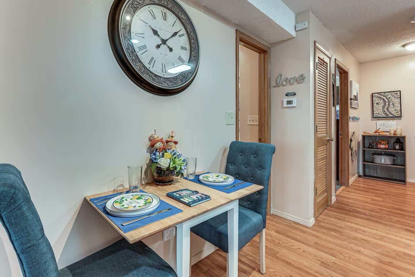 A dining area with a table set for two, a wall clock, and a decorative wall piece.