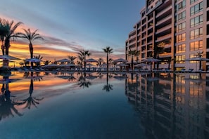 A hotel pool with lounge chairs and umbrellas, surrounded by palm trees and a modern building. - Luxury Ocean Front 2-bedroom Condo in peaceful Cabo San Lucas (Cabo San Lucas)