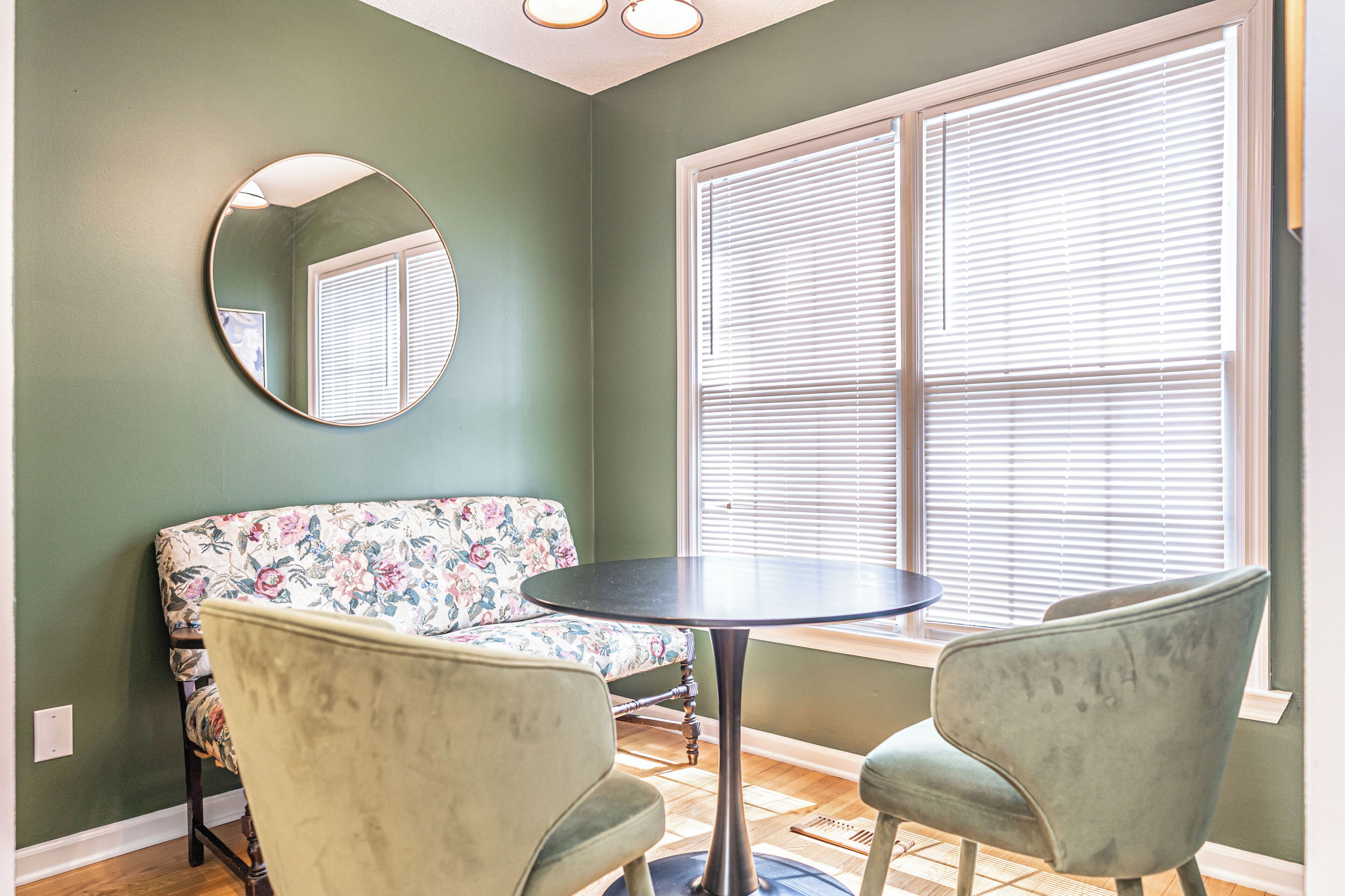Een kamer met een groene muur, een ronde spiegel, een bloemenbank, twee fauteuils, een kleine ronde tafel en een raam met zonwering.