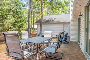 Une terrasse extérieure avec une table et des chaises, entourée d’arbres et avec une maison en arrière-plan.