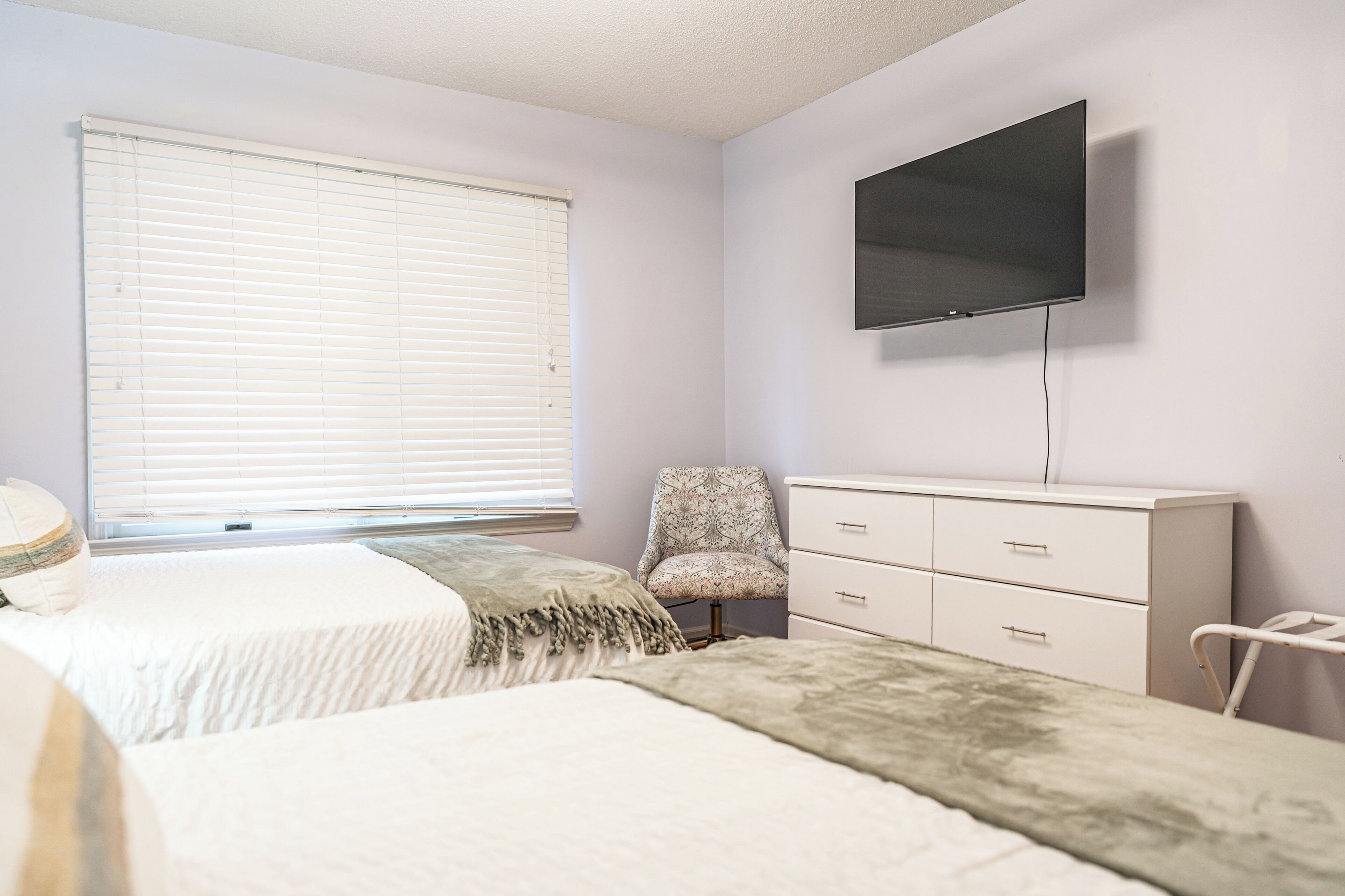 A bedroom with two beds, a chair, a dresser, and a TV mounted on the wall.