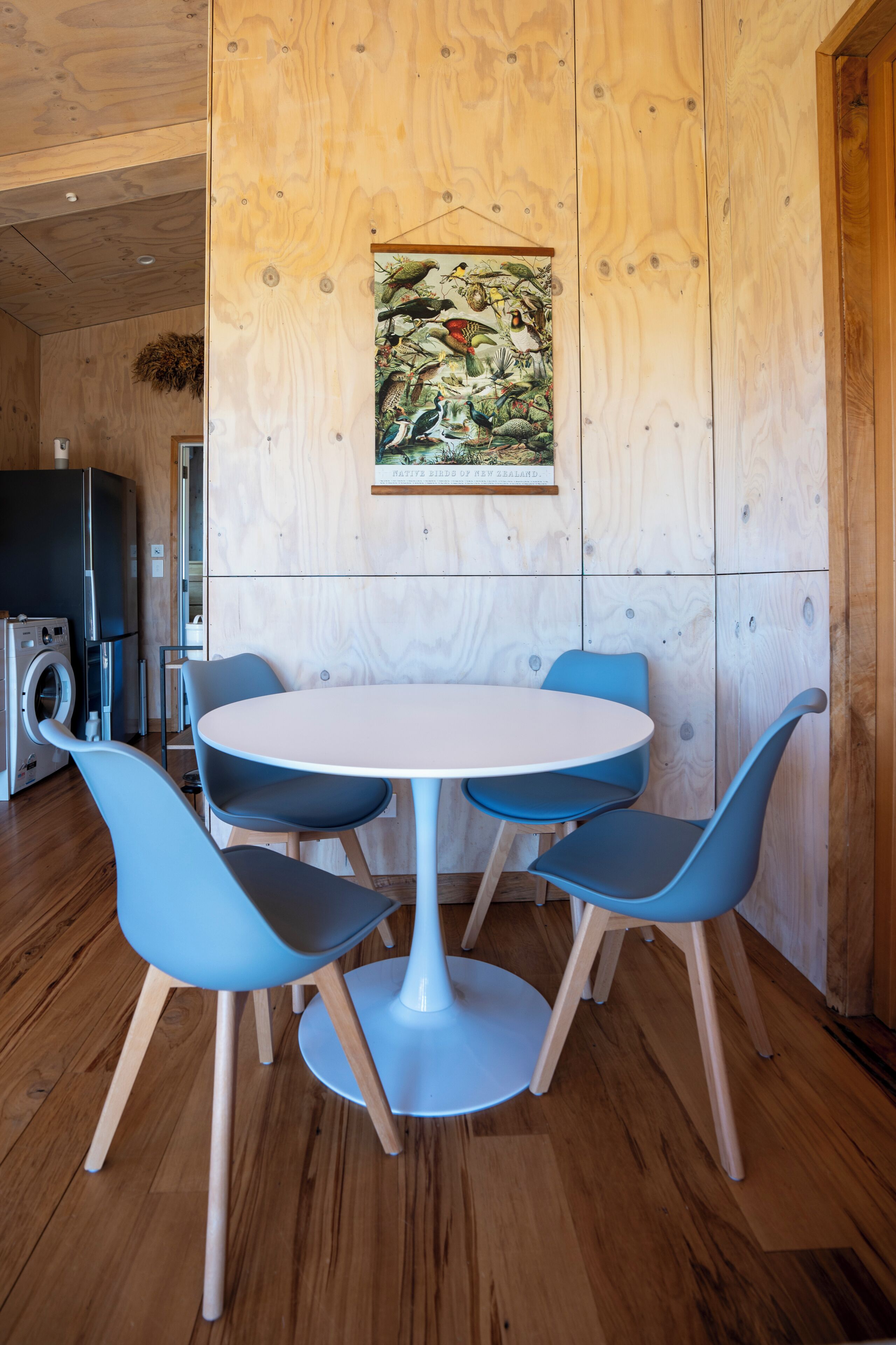 Une cuisine moderne avec une table ronde et des chaises bleues, un mur en bois orné d’une illustration d’oiseau et un lave-linge.