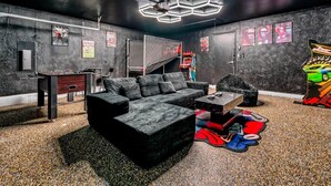 Una habitació amb un sofà seccional negre, una taula petita i una zona de jocs amb taula de futbolí i màquina recreativa.