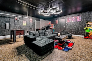 A room with a black sectional sofa, a small table, and a gaming area with a foosball table and arcade machine. - Retro Dallas Gem: Game Room + Fire Pit (Dallas)