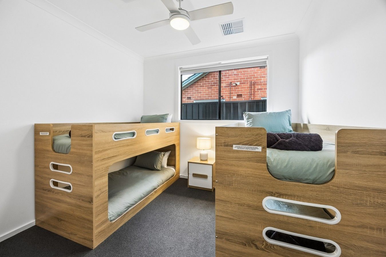 A room with a bunk bed, a desk, a chair, a lamp, and a window with a view of a brick building.